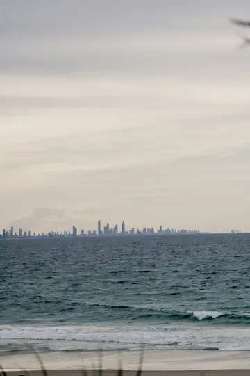 A wide landscape photo showing a distant city skyline with numerous tall buildings across a calm, grey-blue ocean. Gentle…