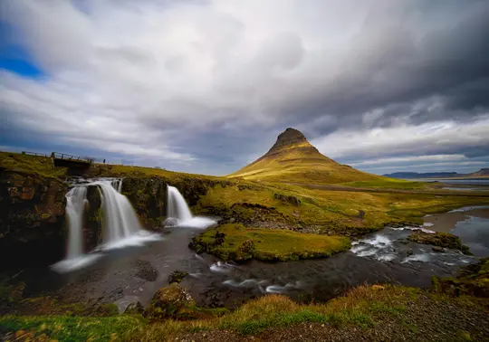 Et bredt landskabsfoto af Kirkjufell-bjerget, en kegleformet tinde, med Kirkjufellsfoss-vandfaldet i forgrunden.