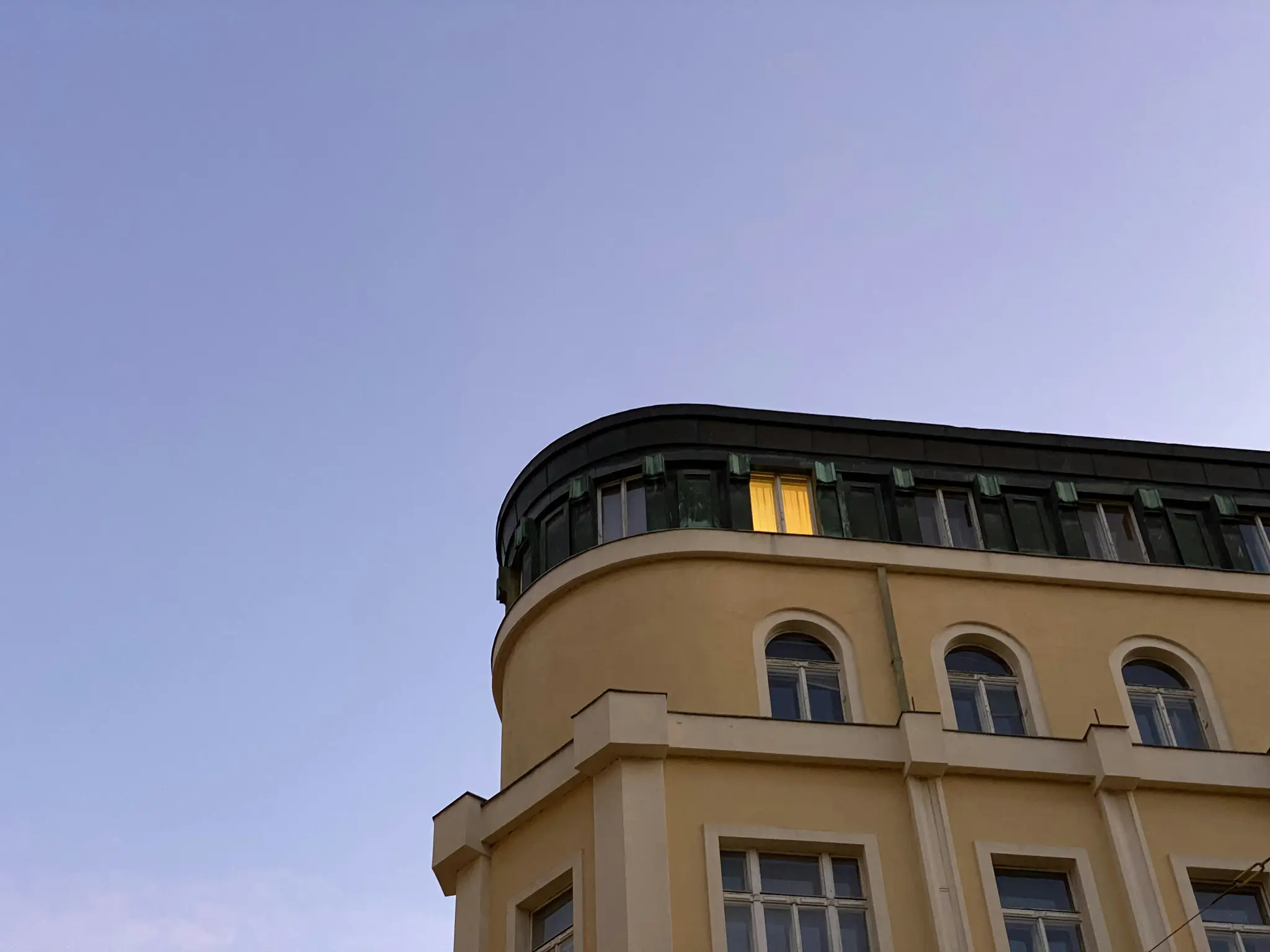 Et lavvinklet billede af et bleg-gult bygningshjørne mod en klar, lyseblå himmel. Et vindue på øverste etage lyser…
