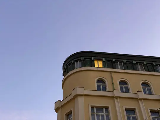 Alacsony szögű felvétel egy halványsárga épületsarkról tiszta, világoskék égbolttal szemben. A legfelső emeleten egy ablak