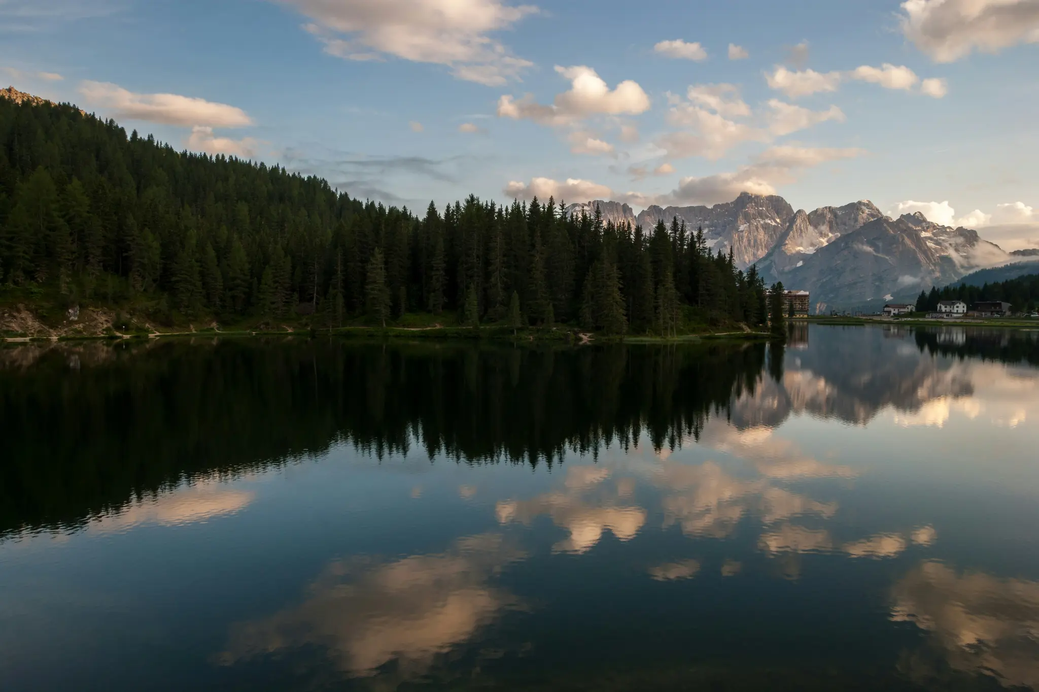 Eine weite Aufnahme eines ruhigen Bergsees, der einen dichten grünen Wald am linken Ufer und schneebedeckte Gipfel unter