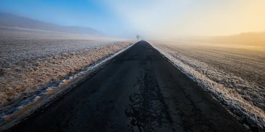 Eine lange, gerade, dunkle Asphaltstraße erstreckt sich in die neblige Ferne, flankiert von Feldern, die mit weißem Reif