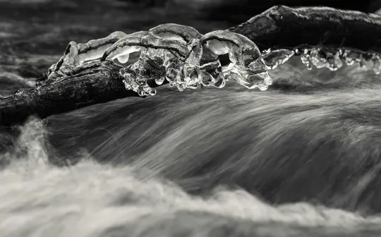 Eine Schwarz-Weiß-Nahaufnahme zeigt einen dunklen, eisbedeckten Ast, der horizontal über verschwommenem, fließendem Wasser