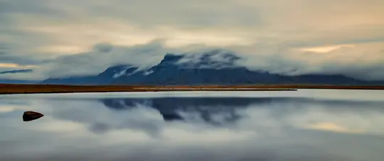 Et bredt landskabsfoto viser et roligt vandområde, der afspejler en bjergkæde, der er delvist skjult af lavthængende skyer.