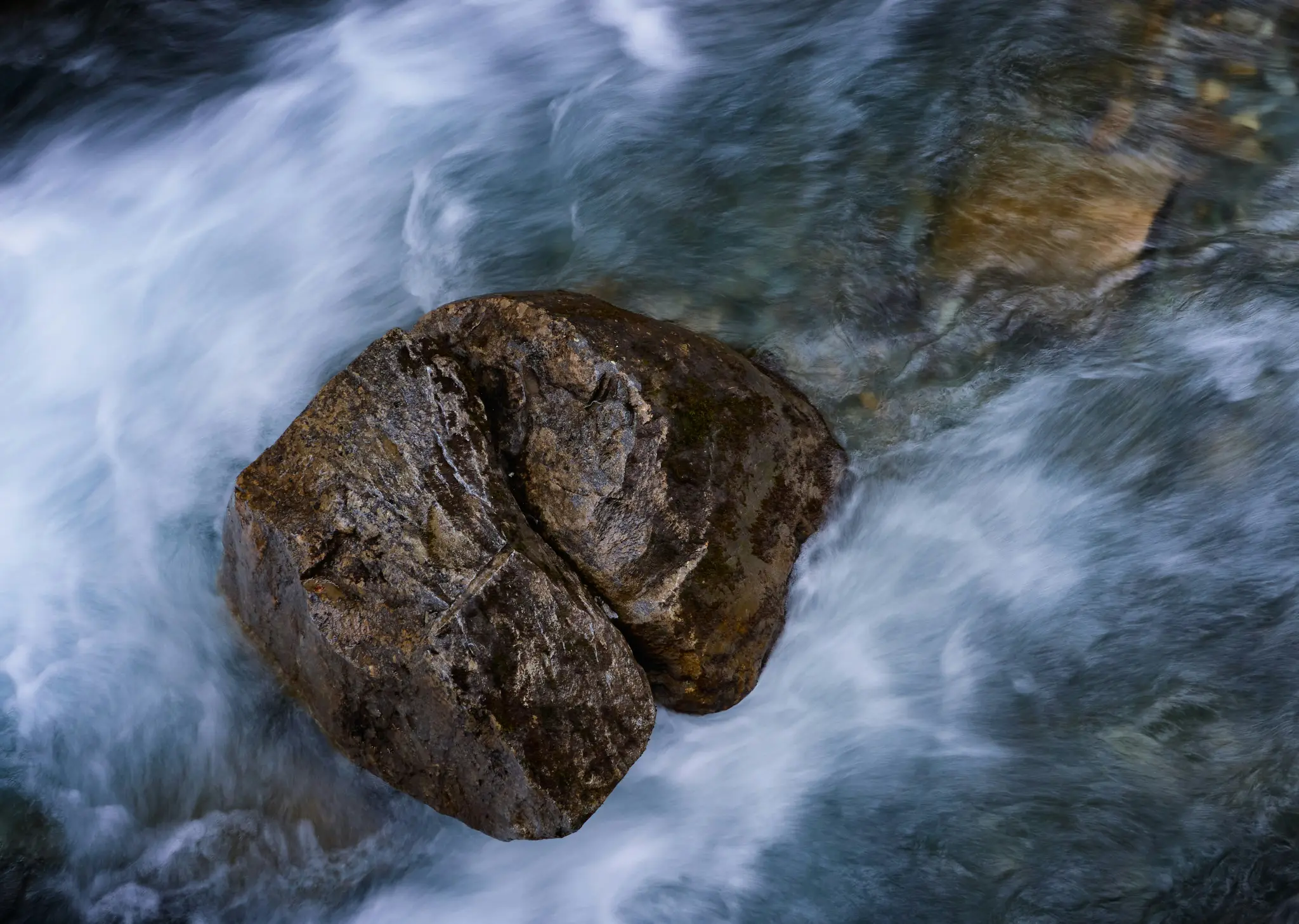 Une vue plongeante d'un gros rocher brun foncé dans un ruisseau. L'eau autour du rocher est floue et blanche à cause du