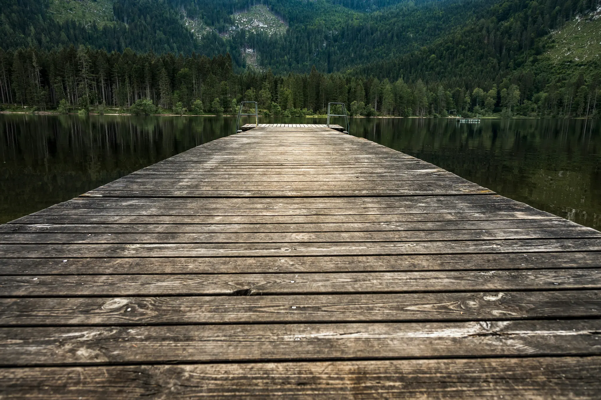 Eine lange, verwitterte Holzpier erstreckt sich in einen stillen See und spiegelt den dunkelgrünen Kiefernwald und die Berge
