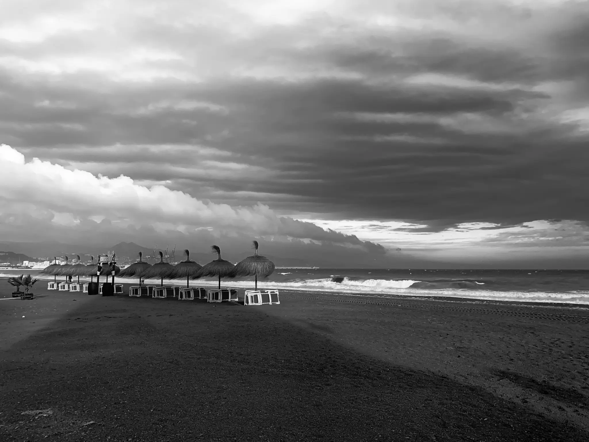Ein Schwarz-Weiß-Foto eines Strandes mit einer Reihe von Strohschirmen und Liegestühlen. Dunkle Wolken füllen den Himmel, und