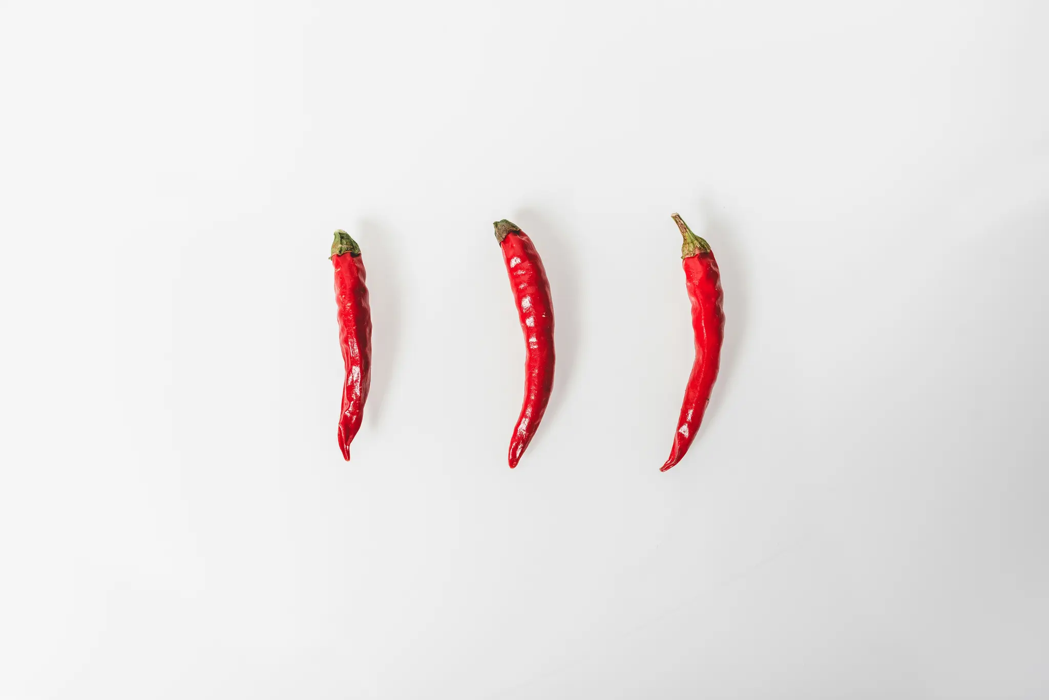 Three vibrant red chili peppers with green stems are arranged vertically and evenly spaced on a stark white background.