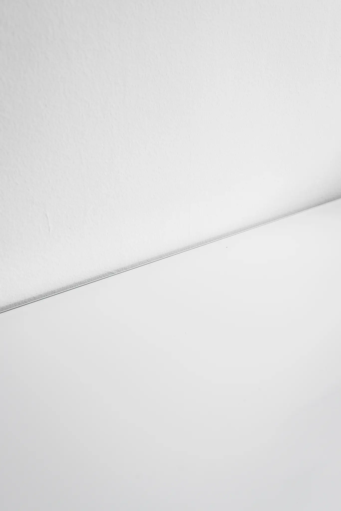 O fotografie minimalistă care arată un perete alb texturat care se întâlnește cu o suprafață albă netedă, separate de o linie