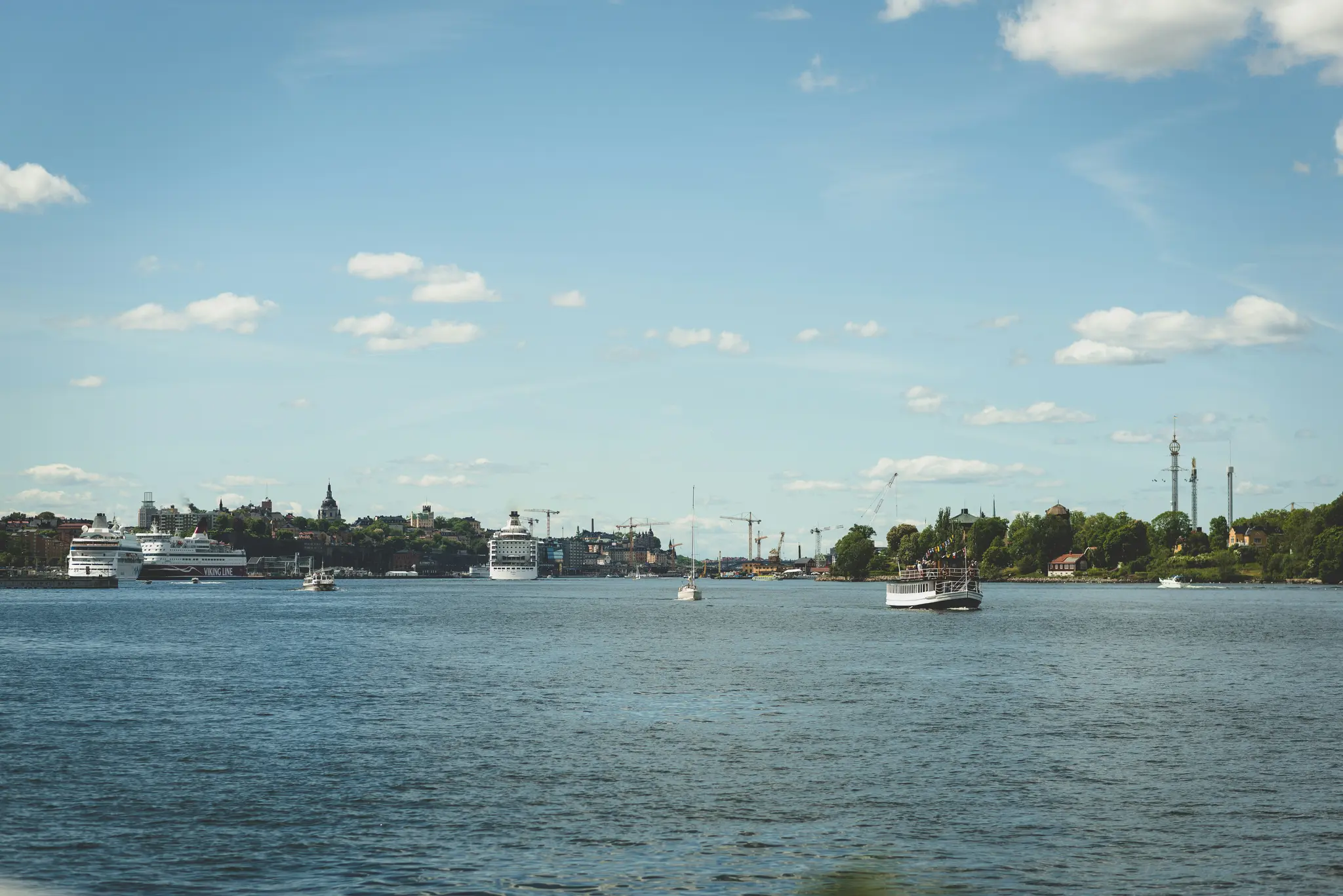 Laaja maisemakuva, joka näyttää vesialueen etualalla ja kaupungin siluetin veneineen etäällä sinisen taivaan alla…