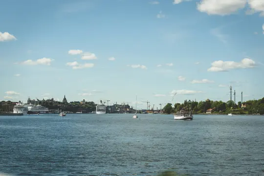 Et bredt landskabsfoto, der viser et vandområde i forgrunden og en by silhuet med både i det fjerne under en blå himmel…