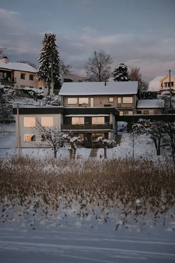A winter landscape showing several snow-covered houses on a hill, with warm light reflecting in their windows. In the…