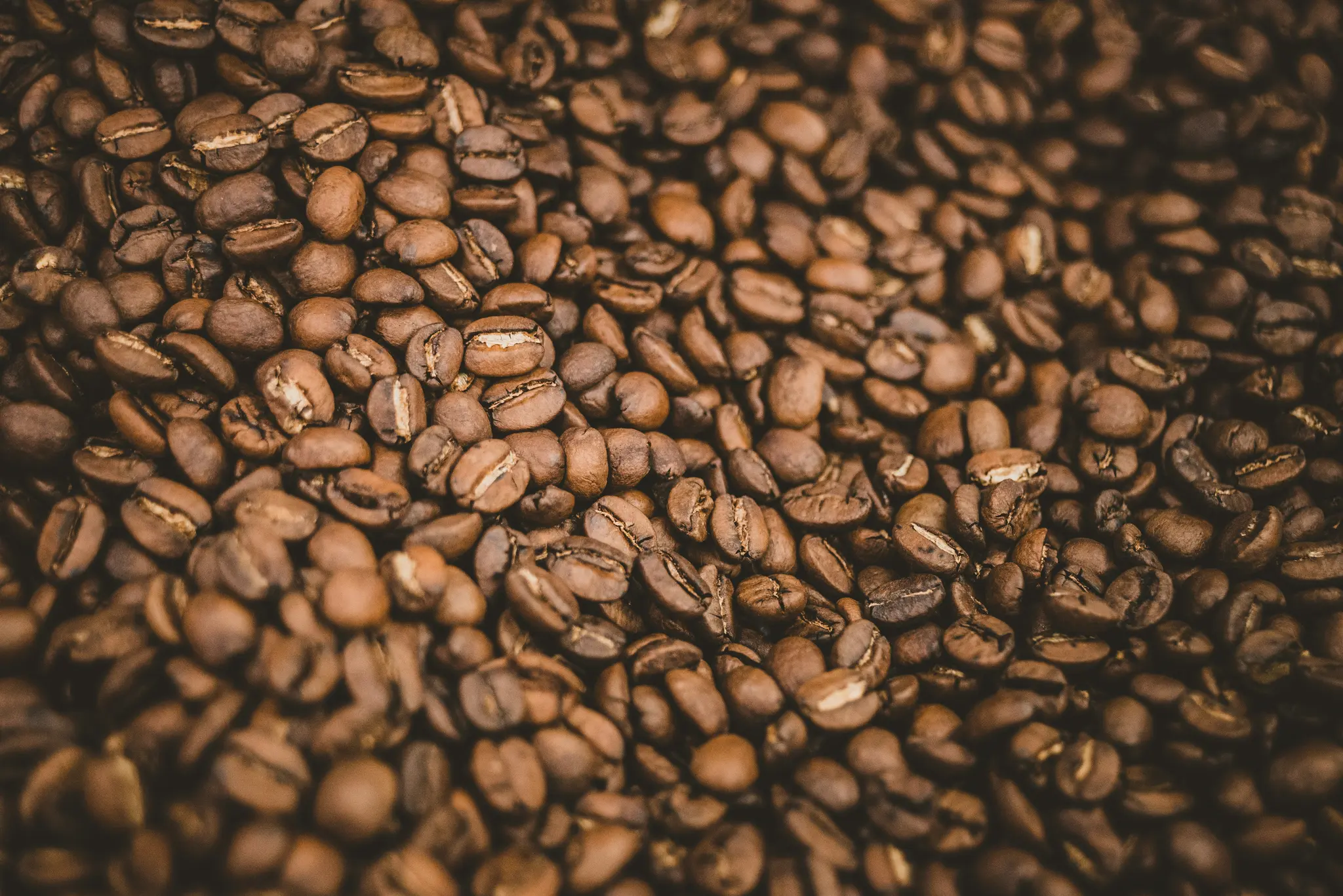 A close-up, overhead shot of numerous roasted coffee beans, filling the entire frame with their dark brown and…