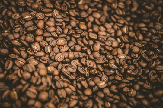 A close-up, overhead shot of numerous roasted coffee beans, filling the entire frame with their dark brown and…