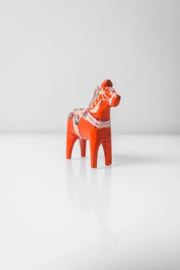 En lille, orange Dala-hestefigur med hvide og blå malede detaljer står på en blank hvid overflade og reflekterer sin…