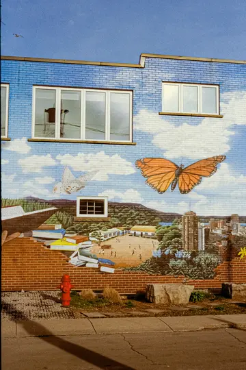 Et levende murale på en murstensbygning viser en blå himmel med skyer, to store sommerfugle, et landskab med bjerge, en…