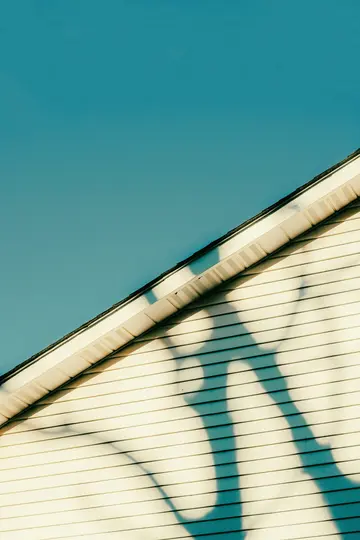 A close-up, abstract photo of light beige vinyl siding on a house, with a clear blue sky above. Dark blue shadows of tree…