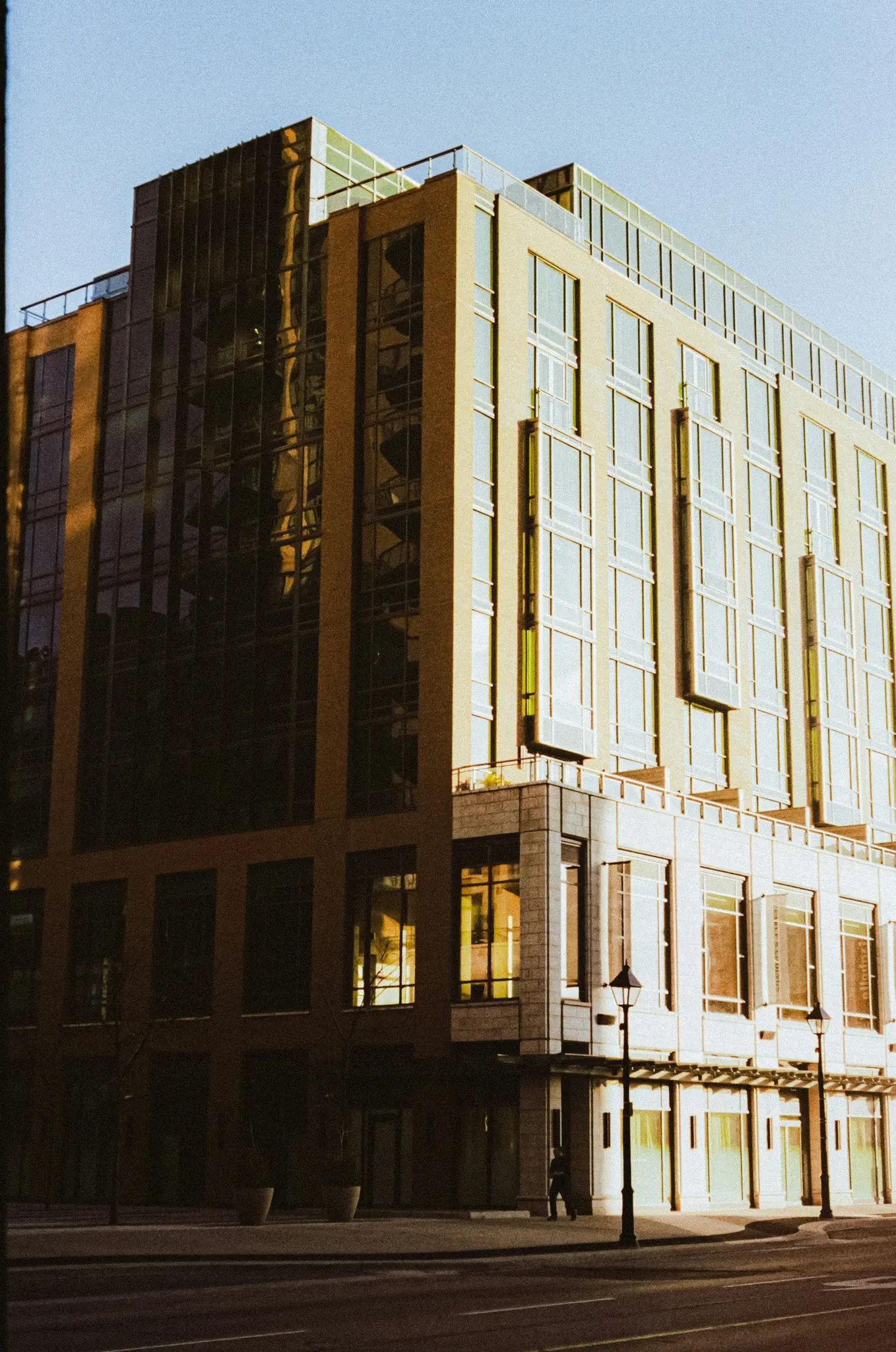 A tall, modern building with a glass and light-colored facade stands at a street corner, bathed in golden hour light.