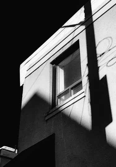 Et sort-hvidt fotografi af en bygningsfacade med høj kontrast. Et vindue er synligt, og stærke, kantede skygger falder hen