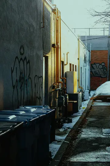 Úzká ulička v zimě se sněhem na zemi. Šedé zdi jsou pokryty graffiti a podél nich vedou žluté trubky.