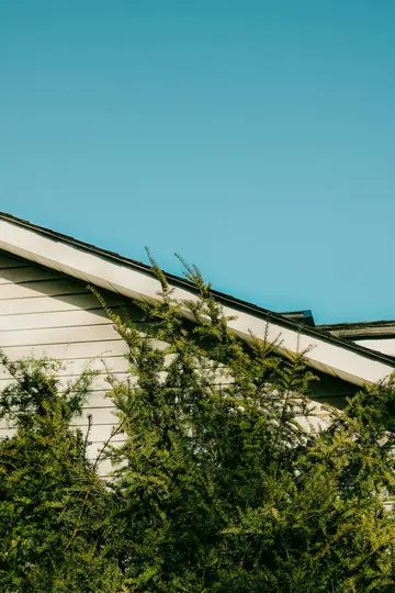 En klar blå himmel dominerer den øverste halvdel af billedet, med husets hvide beklædning og mørke tag nedenunder, delvist…