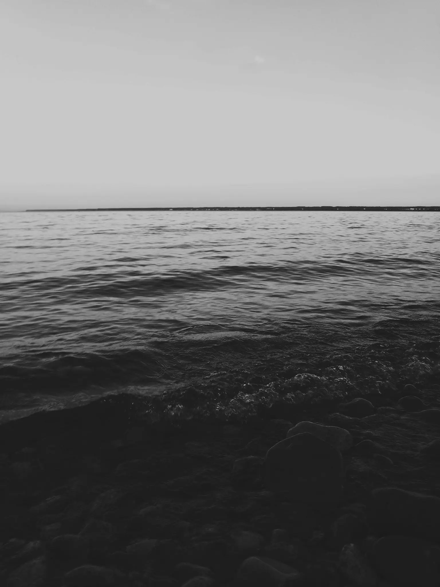Una foto in bianco e nero di acqua calma con piccole increspature, una massa terrestre scura e lontana e un cielo chiaro e