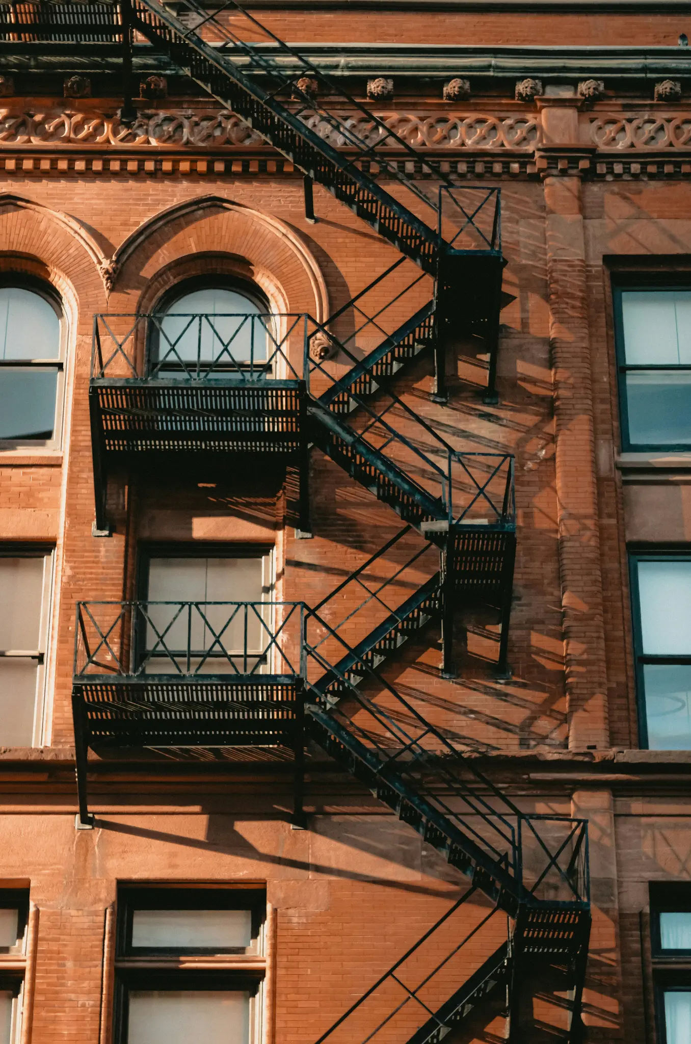 A black metal fire escape with multiple landings and stairs casts shadows on a warm orange-red brick building.