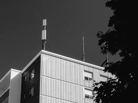A black and white low-angle photo of the top of a building with vertical siding and several antennas on its roof. Dark tree…