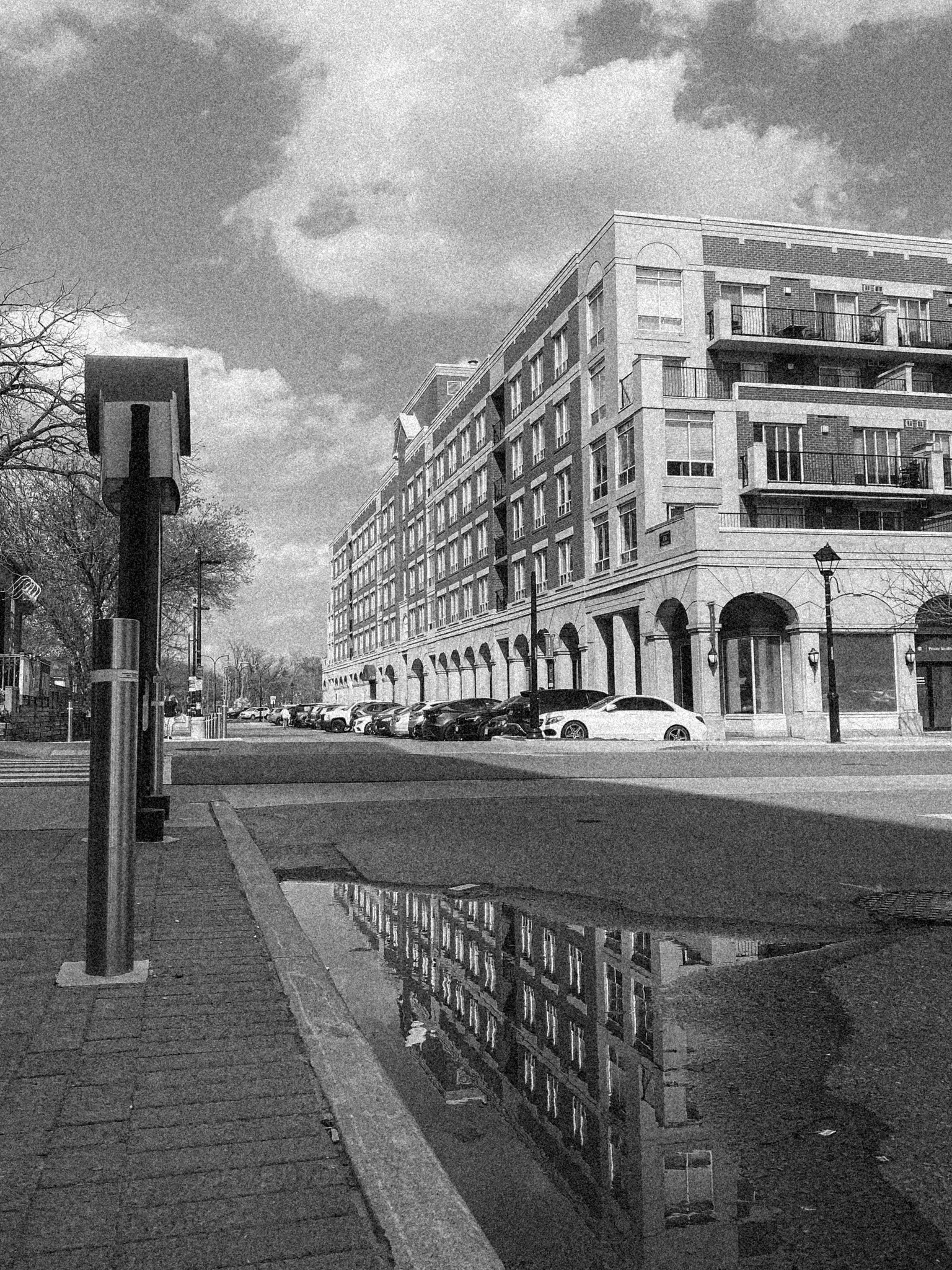 Černobílá fotografie velké budovy s balkony odrážející se v pouliční kaluži. Podél ulice jsou zaparkovaná auta.