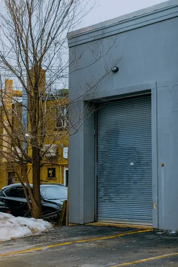 En grå bygning med en stor, lukket, bølget metaldør til garage. Et bart træ står til venstre og delvist skjuler…