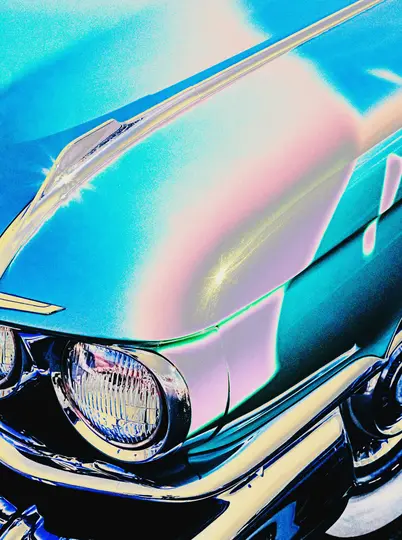 A close-up, high-contrast, and color-shifted image of a vintage car's front, featuring a headlight, chrome, and hood in…