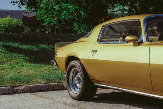 Un coche vintage dorado está aparcado sobre una superficie de asfalto junto a una colina cubierta de hierba con árboles. El