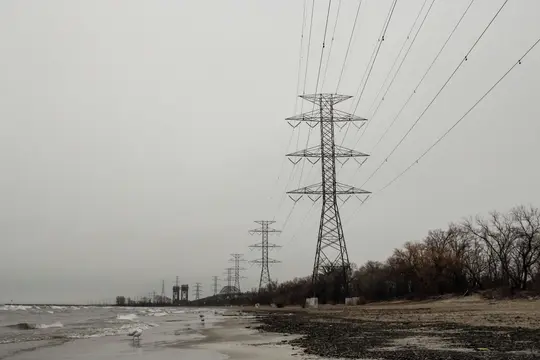 En lang række høje, metaliske elmaster strækker sig ud i det fjerne langs en øde, grå strand med nøgne træer.