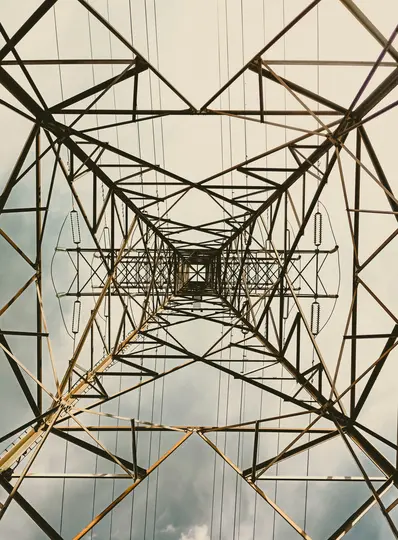 En lavvinklet, opadrettet udsigt ind i den komplekse, geometriske metalramme af en stor elmast mod en overskyet himmel.