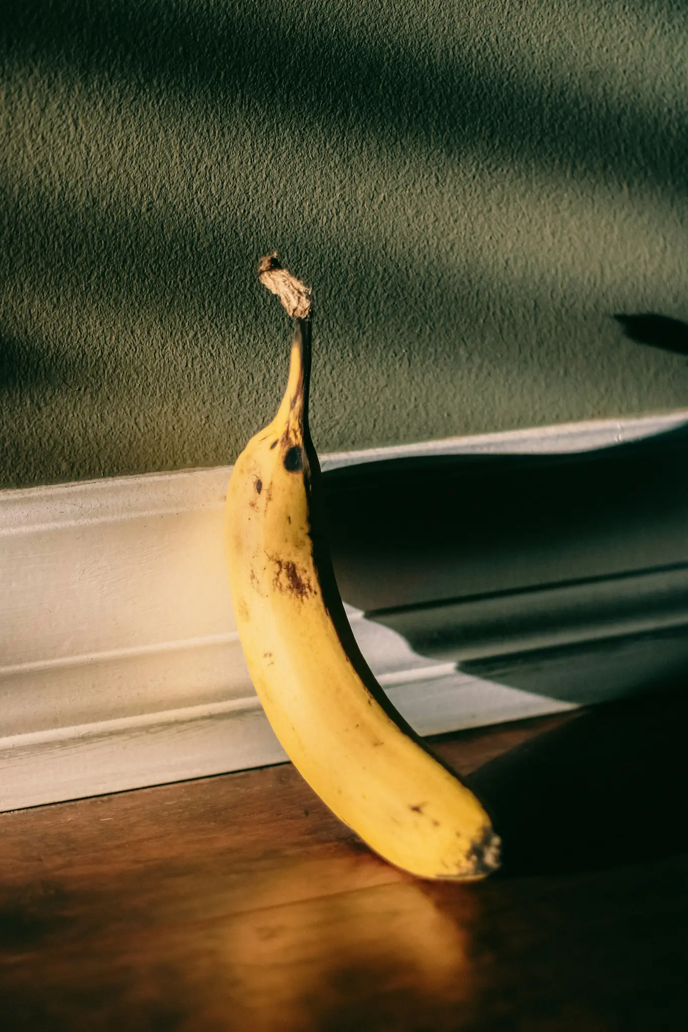 En enkelt gul banan med brune pletter læner sig op ad en tekstureret grøn væg med hvid trim og kaster en lang skygge.