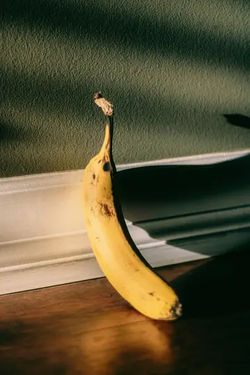 En enkelt gul banan med brune pletter læner sig op ad en tekstureret grøn væg med hvid trim og kaster en lang skygge.