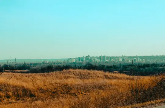 Et bredt landskabsbillede, der viser et felt med tørt, gyldenbrunt græs i forgrunden med en bys skyline i det fjerne.