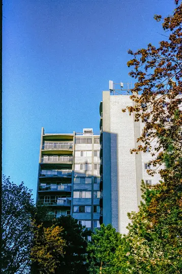 Et lavvinklet billede af to høje, grå beboelsesejendomme med altaner, delvist skjult af grønne og brune efterårstræer.