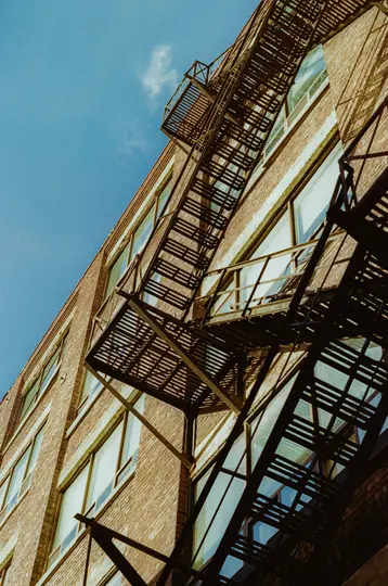 A low-angle shot of a brick building with a dark metal fire escape running diagonally across its facade against a clear…