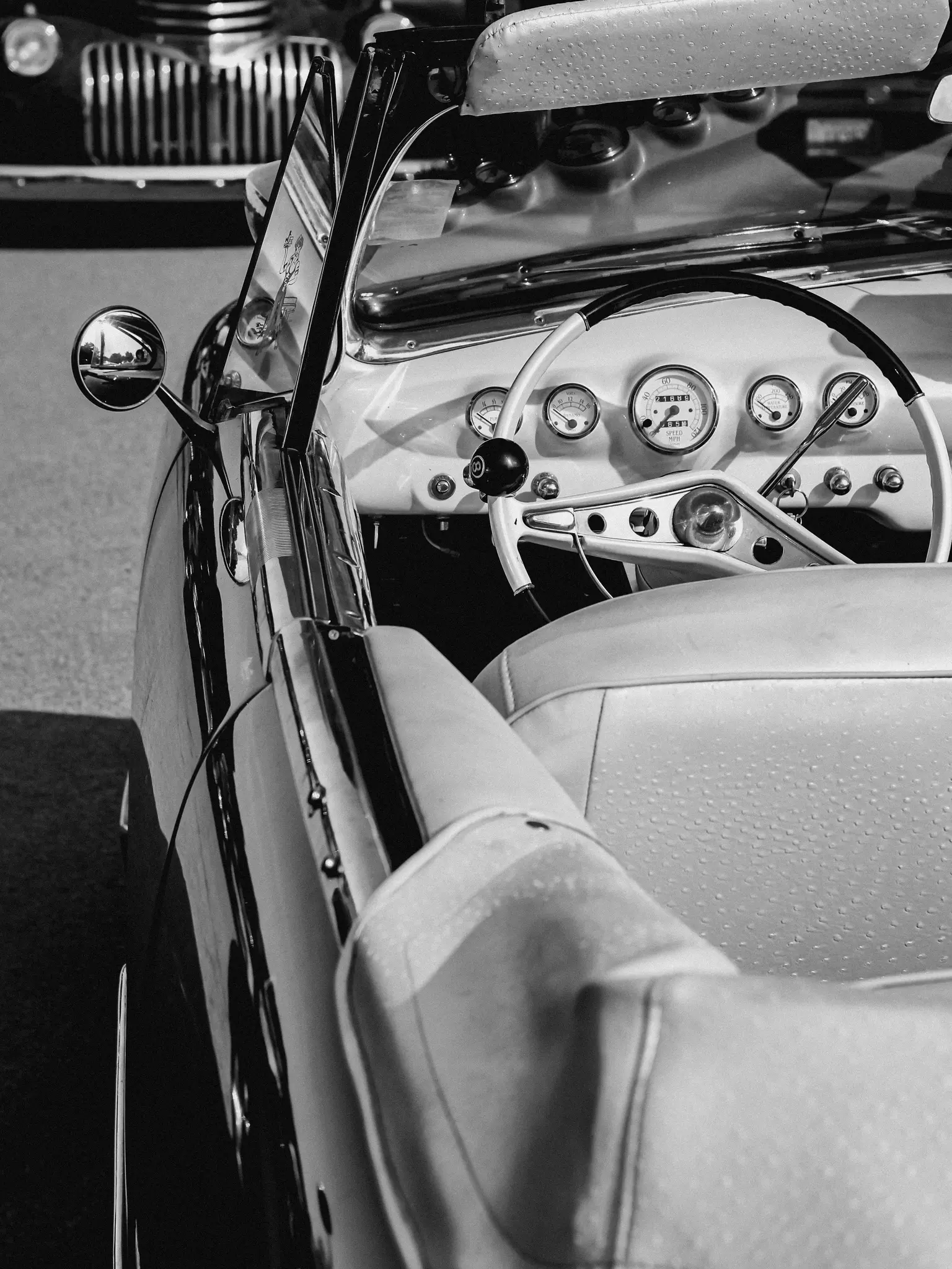 A black and white close-up of a vintage convertible's interior, showing the dashboard, steering wheel, and passenger seat.
