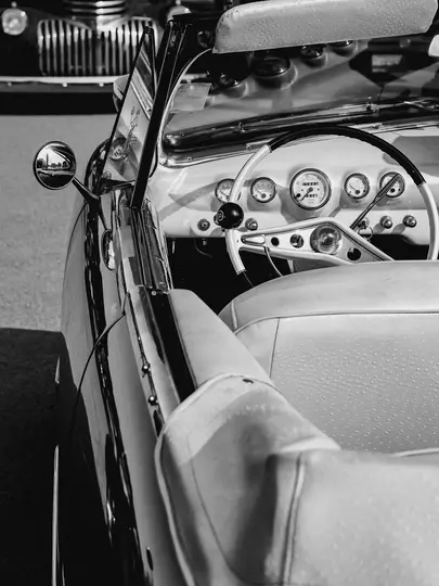 A black and white close-up of a vintage convertible's interior, showing the dashboard, steering wheel, and passenger seat.