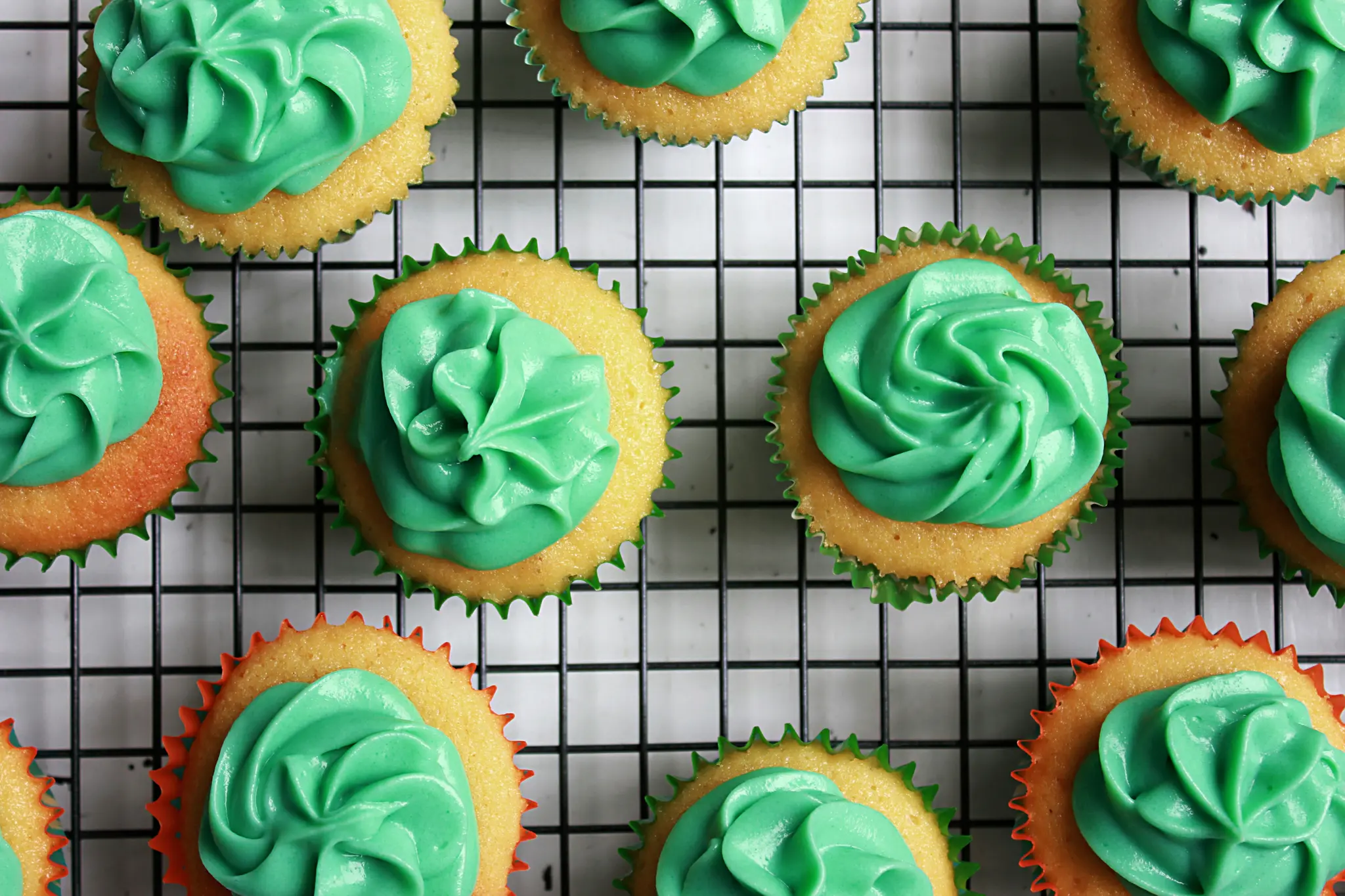 A flat lay photo shows several vanilla cupcakes with bright green swirled frosting, some in green liners, some in orange…