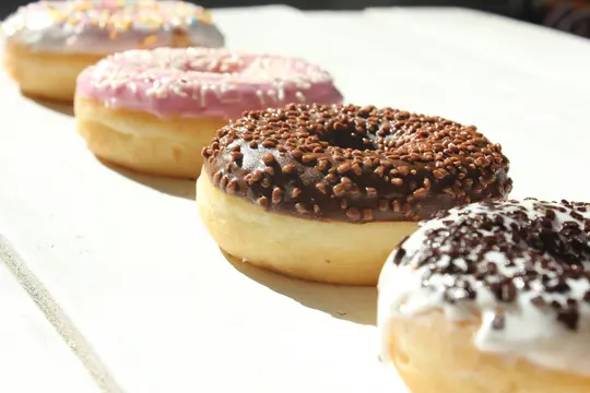 Fire donuts på en diagonal linje. Forfra og bagfra: hvid glasur med mørkt drys, chokoladeglasur med brunt drys…