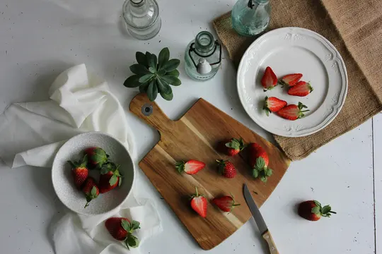 An overhead shot of fresh strawberries, some whole and some halved, on a wooden cutting board, a white plate, and a white…