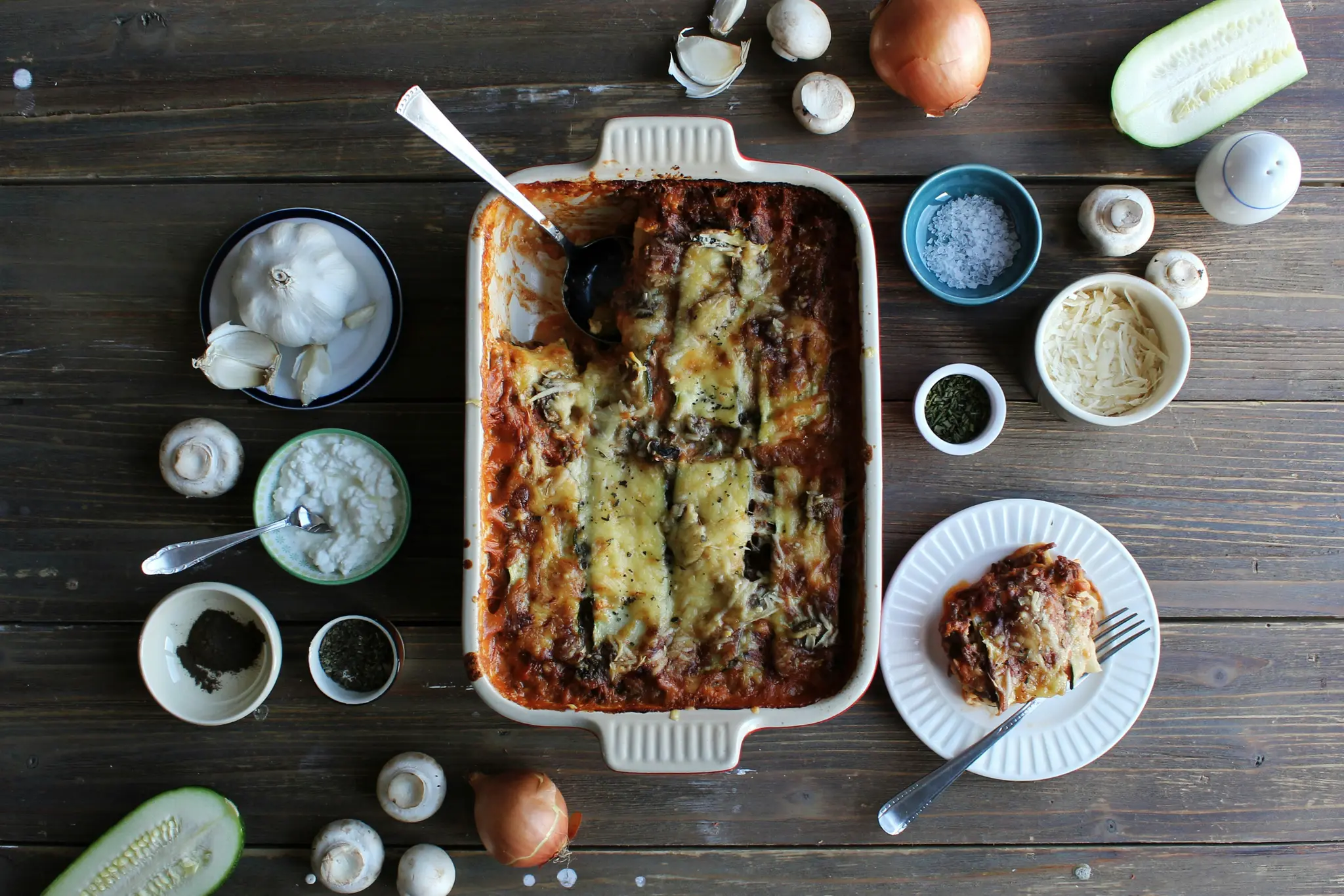 Päältä kuvattu osittain tarjoiltu lasagne uunivuoassa, ympärillään valkosipulia, sieniä, sipulia, kesäkurpitsaa ja...