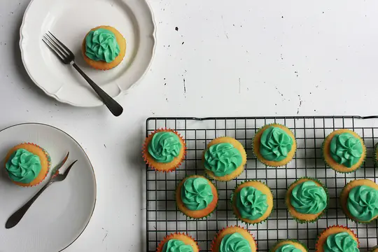 A top-down view of several cupcakes with bright green frosting. Two are on white plates with forks, and many more are on a…