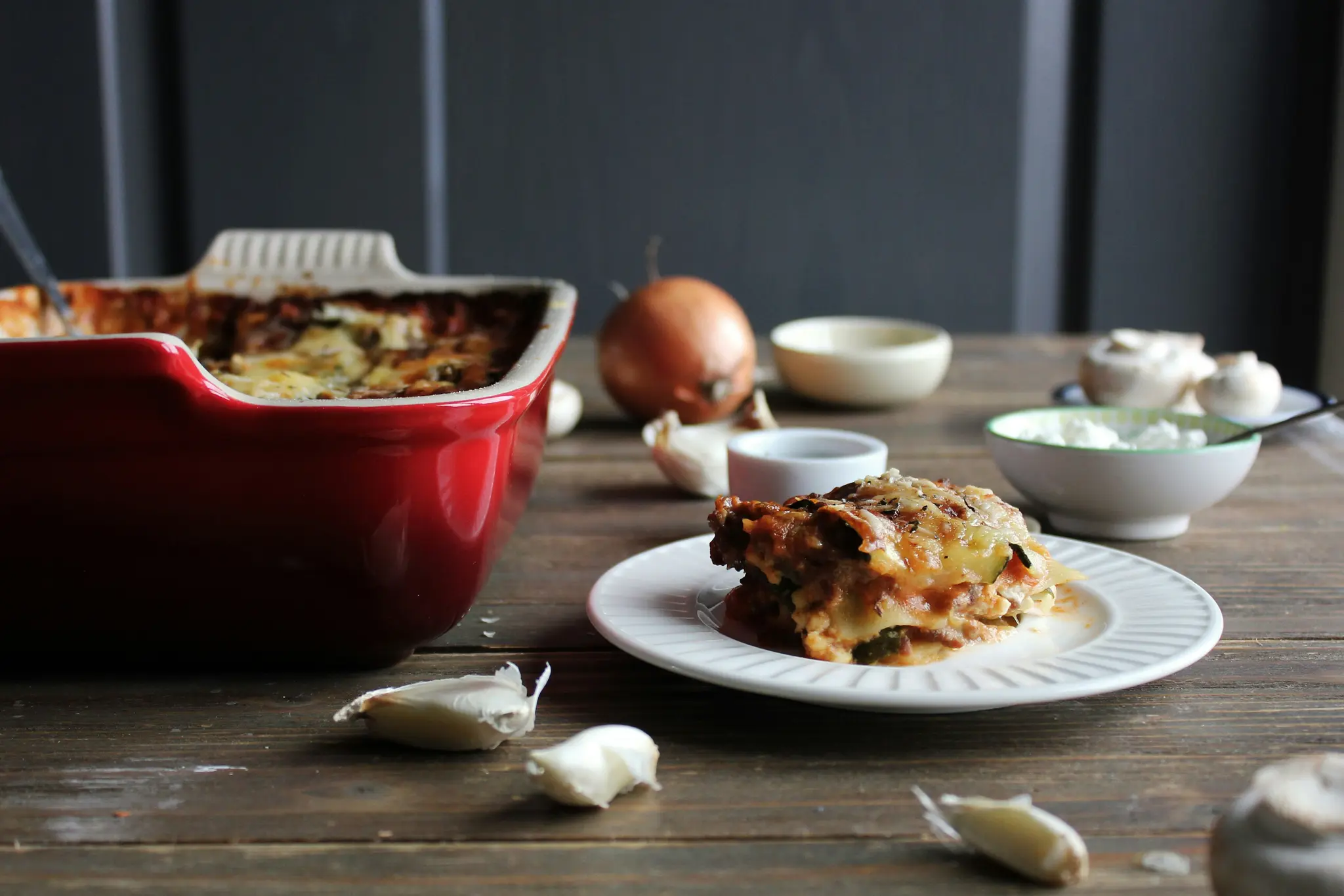 Viipale lasagnea valkoisella lautasella, punainen lasagnevuoka, valkosipulinkynsiä, sipuli ja sieniä puupöydällä…