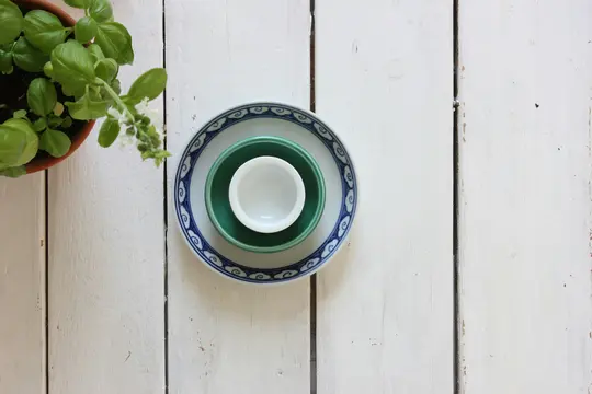 Et ovenfra-billede af en pottebasilikumplante med små hvide blomster og en stak af tre skåle på en hvid træoverflade.