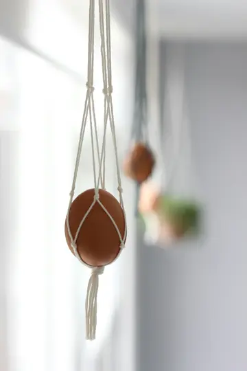 A brown egg is held in a white macrame hanger, hanging in the foreground. The background is blurred with light grey and…