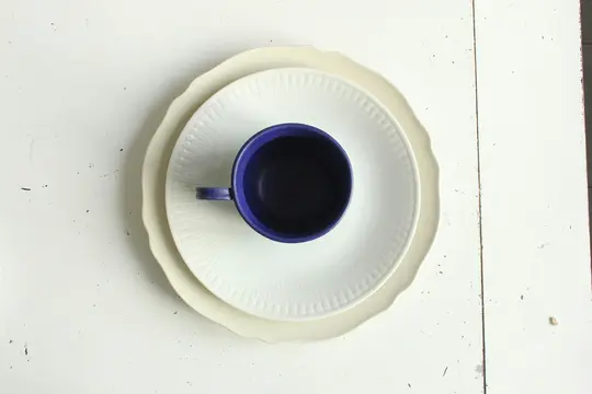 An overhead shot shows a dark blue cup with a handle, centered on a white plate with a ridged pattern. This plate rests on…